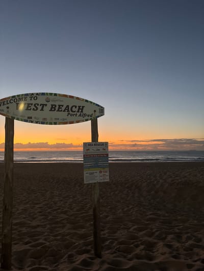 A beach scene at dawn or dusk shows a sign indicating "WELCOME TO WEST BEACH, Port Alfred" with "SUNSHINE COAST" also prominently displayed. The sign is shaped like a surfboard. A secondary sign, labeled "SEA RESCUE" with instructions for escaping a rip current, is attached to the same post. The sky transitions from dark blue to orange, reflecting the sunrise/sunset. The sand is textured, and the ocean's edge is visible in the background.