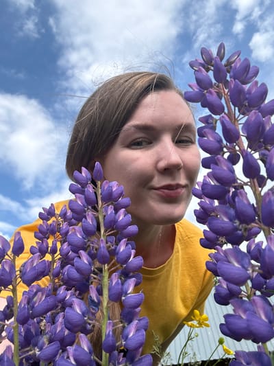 The image shows a woman in a yellow t-shirt peeking through purple lupine flowers against a cloudy blue sky. The perspective is low-angle, with the flowers in the foreground obscuring parts of the woman's face. Her hair is brown and pulled back. The background suggests an outdoor environment, possibly a garden or field. The location is identified as Bolshaya Usa, Russia.