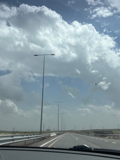 The image shows a highway in the Talin region of Armenia, viewed from inside a moving vehicle. The sky is filled with predominantly gray and white clouds with patches of blue. A straight asphalt road with clear lane markings stretches into the distance, flanked by metal guardrails and grassy areas. Streetlights are evenly spaced along the road. Power transmission towers are visible further away in the distance. The focus is sharpest on the immediate road surface and the nearby guardrails. A portion of the vehicle's interior is visible on the right edge of the frame.