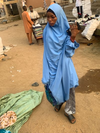 The image depicts an outdoor market scene in Okene, Nigeria, characterized by an unpaved, dusty reddish-brown ground surface. In the foreground, a woman walks from right to left, captured mid-stride. She is wearing a light blue hijab covering her head and body, with a black underscarf visible at the forehead. Her left arm is slightly bent, holding a black smartphone vertically in her hand, with red beaded bracelets visible on her wrist. She wears black sandals and patterned black and white trousers visible beneath her blue garment.
In the mid-ground, to the left of the walking woman, a stack of green fabric lies on the ground, with a smaller bundle of light-colored patterned fabric on top. Further into the mid-ground, on the left, two men are visible. One man, facing the camera's left, wears an orange long-sleeved tunic and matching trousers. The other man, slightly behind him, wears a light-colored shirt and trousers, standing behind a wooden stall displaying neatly folded stacks of multi-colored fabrics.
In the background, on the right side, there is a raised area with several large white sacks and various items, including shoes and clothing, scattered on the ground. A dark, possibly black, object resembling a firearm or tool is partially visible near the base of this raised area. Further back, architectural elements like steps and building structures are discernible, with other blurred figures moving in the distance. The overall scene suggests a busy, open-air retail environment during daylight hours.