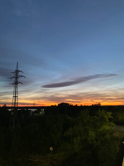 This is a vertical, high-angle shot of a sunset over Novokuybyshevsk, Russia. The sky is filled with a dramatic gradient of colors, from deep blue on the left to vibrant orange and yellow on the right, indicating sunset or twilight. Wispy clouds are scattered across the sky, catching the last rays of the sun. In the foreground, a large, lattice-style electricity pylon stands prominently, silhouetted against the colorful sky. The pylon is supported by wires that extend towards the horizon.
To the right of the pylon, a dense line of dark green trees forms the edge of the landscape, with some lighter green foliage visible. Beyond the trees, faint structures and a distant pylon can be seen. The overall scene evokes a sense of calm and the beauty of nature at dusk, juxtaposed with the industrial element of the electricity pylon. The lighting suggests it is either early morning or late evening. No people are visible in the image. There is a small circular sign with a prohibition symbol on the right side of the image.