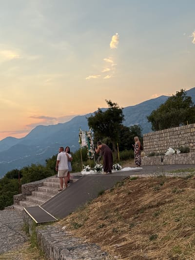 This media file depicts an outdoor scene at sunset in Čelobrdo, Montenegro. The foreground shows a stone wall, overgrown with dry grass and some green vegetation. A dark, wooden walkway runs along the wall, with steps leading down on the left.

Several people are present, appearing to be part of a gathering or ceremony. One woman in a patterned dress stands near a stone wall, looking down and to her right. Further down the walkway, another person in a dark dress is bending over, also near floral decorations. A couple, a man in a white t-shirt and shorts and a woman in a blue top and white shorts, are standing together, with the woman leaning on the man. They appear relaxed and happy.

In the background, a mountain range is visible, with layers of blue and purple hues, suggesting a significant distance. The sky is a gradient of soft orange and blue, indicative of sunset. The overall atmosphere seems serene and pleasant, with the golden light of the setting sun casting a warm glow on the scene. The presence of floral arrangements suggests a special occasion, possibly a wedding or a celebration.