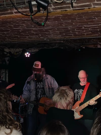 A tilted, low-angle shot captures a band performing indoors. A guitarist in a black t-shirt and a red guitar strap stands at the top. Below, a vocalist/guitarist, wearing a denim jacket, a graphic tee, and a white baseball cap, is mid-performance. A third guitarist and partial audience members appear in the foreground. A brick wall and lighting fixtures are visible on the left.