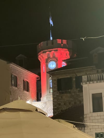 A clock tower in Herceg Novi, Montenegro, is illuminated with red light. The clock face shows the time as approximately 10:11. The tower has crenellated battlements and a flag at its peak. In the foreground, there is a large, pale canopy, likely an umbrella, and parts of stone buildings. The scene is at night, with a dark sky.