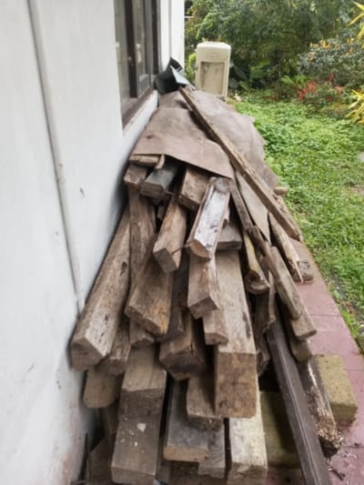 An outdoor scene in Naya Andura, India, displays a large, disorganized pile of weathered wooden planks and logs stacked against a light-colored exterior wall of a building. The wood varies in size, appearing aged and discolored with greyish-brown tones. A dark-framed window is partially visible in the upper left section of the wall. Directly beneath the woodpile and along the wall is a narrow strip of reddish-brown paving or tile. Beyond this paved area, the ground transitions to dense, green ground cover and foliage, extending into the background. Taller green trees are visible in the distance, alongside patches of red flowering plants. A dark, irregular object is situated between the woodpile and the wall near the window. Further back in the green foliage, a light-colored, rectangular object, possibly a discarded appliance, stands upright. No individuals are present in the scene.