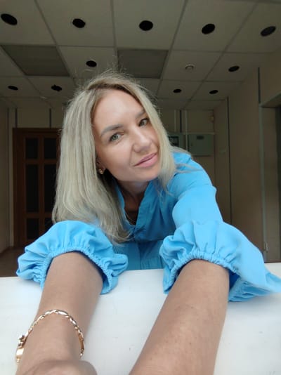 A woman with blonde hair, wearing a blue medical coat, poses for a selfie. Her hair is shoulder-length and slightly wavy. She has light skin and green eyes, and she is smiling slightly. The woman is leaning forward, her arms resting on a white surface. She is wearing a gold bracelet on her left wrist. The background is a room with a drop ceiling, light-colored walls, and a wooden door. The setting appears to be an office or medical facility. The image was taken indoors.