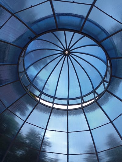 This is a low-angle, close-up photograph looking up at the interior of a blue, ribbed polycarbonate dome structure. The dome is supported by a dark metal framework, which divides the dome into geometric sections. The ribbed texture of the polycarbonate creates a visual pattern of lines. In the center of the dome, a small circular opening is visible, allowing some light to enter. The structure appears to be an outdoor canopy or pavilion, possibly located in a park or recreational area in Goryachiy Klyuch, Russia, as suggested by the reflections of trees and sky visible through the translucent material. There are no people or animals present in the image. The overall impression is one of architectural design and the play of light through translucent materials.