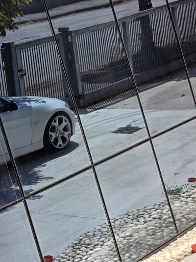A white BMW is parked in front of a modern building with large glass panels. The glass panels reflect a fence, a road, and trees. The scene appears to be a sunny day. The BMW is a coupe with a sleek design and alloy wheels. The fence is made of metal bars and is painted gray. The road is asphalt and has tire marks on it. The trees are green and leafy. The image is taken from a low angle, looking up at the building. The lighting is bright and there are strong shadows. The location is Stanghella, Italy.