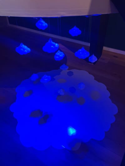 The image displays an interior art installation located in Sélestat, France, predominantly illuminated by blue light. From an overhead, off-white rectangular structure, approximately nine translucent, cloud-shaped objects are suspended at varying heights by thin, clear filaments, each emitting internal blue luminescence. Below, a large, irregularly contoured white surface, approximately two meters in its longest dimension, rests on a light-toned wooden floor. This surface is intensely illuminated by blue light, rendering it luminous. Scattered upon this glowing surface are multiple smaller, opaque, cloud-shaped forms and various dark, irregular silhouettes. A concentrated bright spot of blue-green light emanates from one area of the large illuminated surface. The surrounding background consists of dark-toned walls and the continuation of the wooden floor, with a dark, vertical structural element visible on the right side of the frame. No individuals are present in the scene.