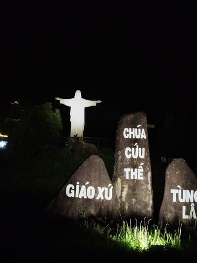 The image presents a nighttime view of a religious monument in Lạc Dương, Vietnam. A large, white statue of Jesus Christ with outstretched arms stands prominently on a raised platform or hill, illuminated against a dark, starless sky. Dark foliage and trees are visible to the left of the statue.
In the immediate foreground, three massive, dark rocks or boulders are artificially lit, contrasting sharply with the darkness. Each rock features white Vietnamese text painted on its surface. The rock on the left displays "GIÁO XỨ" (meaning "Parish"). The central and largest rock reads "CHÚA CỨU THẾ" (meaning "Christ the Savior" or "Lord Savior"). The rock on the right, partially visible, shows "TÙNG LÂ," which is likely part of "TÙNG LÂM," possibly referring to a specific parish or location named Tùng Lâm. Green grass around the base of these rocks is also highlighted by the artificial lighting, adding a touch of verdancy to the otherwise dark setting.
The scene is static, capturing the monument in a solemn and reverent atmosphere. The bright illumination of the statue and the inscribed rocks against the deep black of the night sky emphasizes their significance. The image conveys a sense of peace and spiritual presence, typical of a religious landmark after dark.