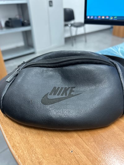 A close-up, eye-level, horizontal shot shows a black Nike fanny pack lying on a wooden desk. The fanny pack is positioned diagonally from the lower left to the upper right of the frame. The Nike logo is prominently displayed in black on the front of the fanny pack. The zipper pull on the left side of the fanny pack has the word "TRAVEL" inscribed on it. The background is blurred, showing office furniture, including shelves with items on them, a chair, and a computer monitor displaying a colorful screen. The overall lighting is neutral, suggesting an indoor environment. The image does not provide any explicit clues to its location within the city of Kamianske, Ukraine.