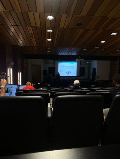 The image depicts a dimly lit lecture hall or auditorium in Lawrence Township, United States, viewed from the rear audience perspective. The ceiling is constructed from horizontal wooden slats with embedded circular downlights. At the front of the room, a large white projector screen displays a slide titled "Seawater," detailing its composition and properties, along with a multi-segmented circular graphic. One individual, the presenter, stands to the left of the screen.
Multiple attendees are seated in rows of black upholstered chairs. In the left foreground, an individual holds a smartphone vertically in their left hand, while their right hand rests near an open laptop displaying a webpage resembling YouTube content. This person wears multiple beaded bracelets on their left wrist. Further forward, a person wearing a red shirt and another wearing a white baseball cap backward are visible, among other partially obscured individuals, many of whom are utilizing laptops. The scene captures an educational presentation in progress.