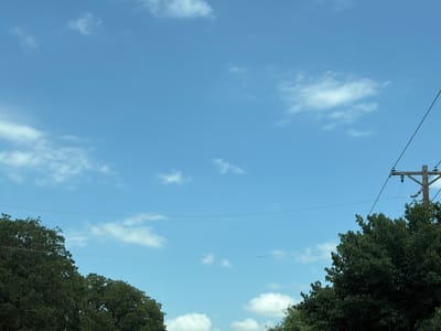 The image is a daytime, medium-angle shot of a bright blue sky with scattered white clouds. In the foreground, the tops of lush green trees frame the bottom corners of the image, with the left side dominated by a dense canopy of broad leaves and the right side featuring a slightly sparser arrangement of foliage. A wooden utility pole with electrical wires stretching across it is visible on the far right. Two horizontal wires are also stretched across the sky. The scene suggests an outdoor environment, potentially a residential area or a park, given the presence of mature trees and utility infrastructure. The location is identified as Grapevine, United States of America. There are no people or animals visible in the image.