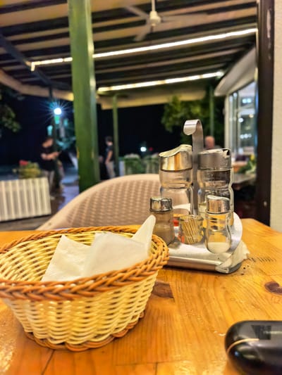 This is a medium shot, eye-level, slightly blurry, outdoor evening photo taken at a restaurant in Velika Plana Municipality, Serbia. In the foreground, a woven basket with white napkins sits on a wooden table. To the right of the basket is a salt and pepper shaker set with a metal handle, a toothpick holder filled with crackers, and a small container of what appears to be milk or cream. The background shows an outdoor seating area with tables and chairs, illuminated by string lights and a bright light fixture. Two blurry figures are visible in the distance. The overall atmosphere suggests a casual dining experience at an open-air establishment.