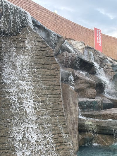 This image shows a close-up view of a man-made waterfall and rock formation in front of a brick building. A red banner with white lettering, reading "PLAZA CARONÍ", is visible on the building. The waterfall cascades down the artificial rocks into a pool of water at the bottom. The scene appears to be an outdoor public space in Guayana City, Venezuela, likely a shopping center or park area given the plaza signage. The sky is overcast.