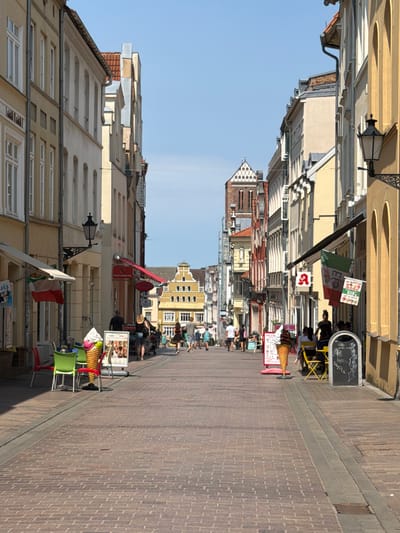 The image depicts a street scene in Wismar, Germany, likely during daytime under a clear blue sky. The street is paved with brick and lined with historic, multi-story buildings on both sides. The architecture features a mix of classical and traditional European styles, with various facade colors including yellow, beige, and red.
On the left side of the street, there are awnings over shop fronts and outdoor seating areas with green and red chairs. A large, colorful ice cream cone sculpture is prominently displayed near the sidewalk, accompanied by a sign advertising ice cream with images of cones. Further down the street on the left, people are walking and seated at tables.
On the right side of the street, more awnings are visible, along with a black lamp post attached to a building. A pharmacy sign with a red 'A' symbol is displayed. An Italian flag is hanging from a pole. Other signs and advertisements are also present. People are walking along the street and seated at tables outside establishments. In the background, a tall building with a distinctive stepped gable roof is visible, and further in the distance, a church tower with a checkered facade can be seen. The overall atmosphere appears to be that of a lively pedestrian zone in a historic town.