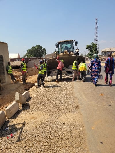 The image depicts a daytime road construction scene in Ube, Nigeria, under a clear blue sky. In the midground, a large yellow Caterpillar bulldozer is positioned with its front blade lowered, containing several white blocks. Six male workers are present around the bulldozer and the immediate construction area, mostly wearing high-visibility vests (yellow or green) and some with hard hats. One worker in a red shirt and red hard hat stands near the bulldozer's blade, with his right arm raised. Another worker in a blue cap and blue vest holds a red object. One worker leans against a light-colored building wall on the far left.
The foreground on the left features a section of ground covered with light brown gravel, bordered by several concrete curbstones, some upright and some toppled. To the right, an existing darker asphalt or dirt road surface is visible. Two women, wearing colorful patterned traditional clothing, walk away from the camera on the right side of this road.
The background includes various low-rise buildings with corrugated roofs, scattered green trees, and a tall, slender cellular communication tower prominently positioned on the right side. The overall scene suggests ongoing infrastructure development.