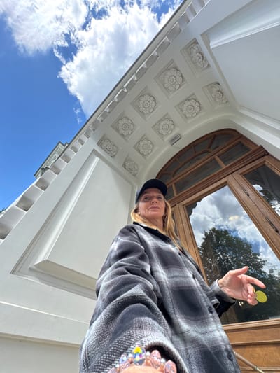 A woman wearing a black baseball cap and a grey and black plaid jacket stands in front of a building with ornate white trim and a large wooden door with glass panes. The building has a neoclassical architectural style, characterized by the decorative floral motifs on the ceiling and the fluted columns. The sky is blue with scattered white clouds, reflecting in the glass of the door. The woman is looking up and to the left, with her right hand outstretched, holding a small yellow object. Her left hand is visible at the bottom of the frame, holding what appears to be a collection of colorful, beaded rings. The image is taken from a low angle, emphasizing the grandeur of the building. The location is Sinyavino, Russia.