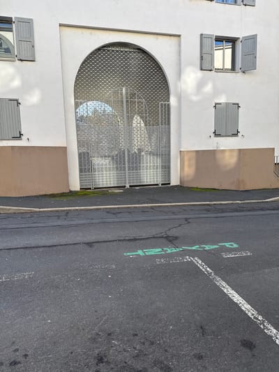 This image captures a street-level view of a building facade and adjacent asphalt road in Le Puy-en-Velay, France. The building presents a white upper section and a beige lower foundation. Central to the facade is a large arched entrance, secured by a partially lowered metal roll-down grille and a double-leaf metal gate with a square grid pattern. Four windows with light-gray wooden shutters are visible: two on the upper left are closed, one on the upper right is open revealing a reflective pane, and one on the lower right is closed. A small amount of moss grows on the sidewalk at the base of the arched entrance. The foreground consists of an asphalt road with visible cracks. Green stenciled text reading "PAYSAGE" is visible on the asphalt, along with a faded white parking line marking. A concrete curb delineates the sidewalk from the road. No individuals, vehicles, or animals are present. Distinct shadows are cast on the building's white facade, indicating directional sunlight.