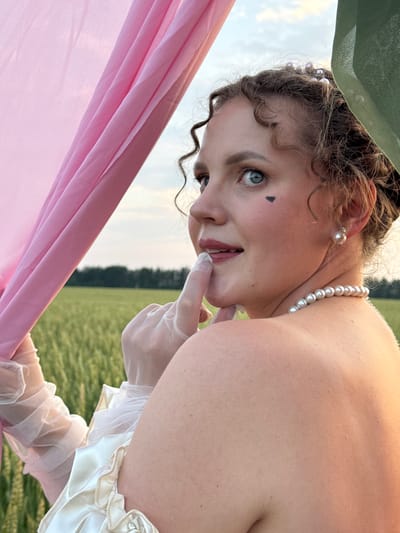 A young Caucasian woman with curly brown hair is in a field of wheat, partially obscured by sheer pink and green fabric. She is looking over her shoulder towards the camera, with her right index finger touching her lips. She has a small black heart drawn on her left cheek, and she is wearing pearl earrings and a pearl necklace. Her dress appears to be made of white satin with ruffled sleeves. The sky is a pale blue with scattered clouds, and the sun is setting, casting a warm glow on the scene. The location is Foki, Russia.