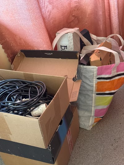 The image depicts an indoor scene, likely a storage area or a room undergoing tidying, with several cardboard boxes and a colorful tote bag filled with items. In the foreground, a large cardboard box is open, revealing a tangled mass of black electrical cords and at least one white plug or adapter. To the right of this box, another open cardboard box contains more miscellaneous items, including smaller cardboard pieces, and a glimpse of a striped fabric bag. Further back and to the right, a large, colorful tote bag with horizontal stripes of pink, orange, yellow, and black, is visible, also containing various objects. Behind the boxes and bag, a textured, pinkish-red fabric, possibly a curtain or draped material, hangs in the background. The floor appears to be carpeted with a speckled grey and beige pattern. The overall impression is one of accumulated belongings. There are no people visible in the image, and no specific landmarks or contextual clues suggest the city of Naro-Fominsk, Russia, or any other specific location.