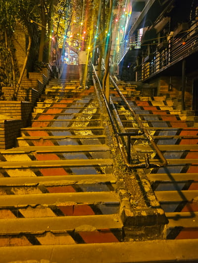 In Beyoğlu, Turkey, a well-lit stairway is illuminated by colorful string lights. The steps are painted with a repeating pattern of yellow, red, and blue triangles. A person wearing a light-colored shirt and dark pants stands at the top of the stairs with their arms outstretched. Buildings line the right side of the staircase, with balconies and windows visible. Trees with dense foliage grow on the left side of the stairs. The scene is captured at night, with artificial lighting casting shadows and highlighting the vibrant colors of the steps.