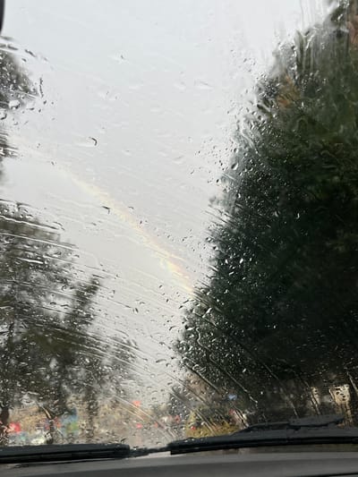 The image depicts a rainy outdoor scene viewed from inside a vehicle, specifically through its water-streaked windshield. Numerous raindrops of varying sizes are visible across the entire glass surface, with some streaking patterns. The sky overhead appears bright and overcast. To the left and right, large, dark green trees are present, their forms blurred by the rain. In the lower portion of the image, the vehicle's black dashboard and two black windshield wiper blades are visible in their resting position. Through the distorted view of the wet windshield, indistinct red lights and other muted shapes, possibly indicating other vehicles or structures, are present in the middle distance. A faint, narrow spectrum of colors, resembling a partial rainbow or light refraction, is visible diagonally in the upper-middle-left area of the sky. The location is Al-Qanawat Municipality, Syria.