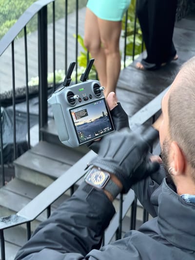 In this image, a person is holding a drone controller, focused on the screen displaying a landscape view with a sunset or sunrise. The person is wearing black gloves and a dark jacket. A smartwatch is visible on their left wrist. The background shows hints of a tropical setting with lush greenery, possibly suggesting Kintamani, Indonesia. Other people are partially visible in the background, with one person wearing a mint green top. The scene appears to be outdoors, possibly on a balcony or elevated area given the railing and stairs. The text on the controller screen indicates information about flight height, altitude, and battery status, suggesting an impending drone flight. The overall mood is one of anticipation and focus on operating the drone. The time of day is likely dawn or dusk, judging by the lighting on the screen.