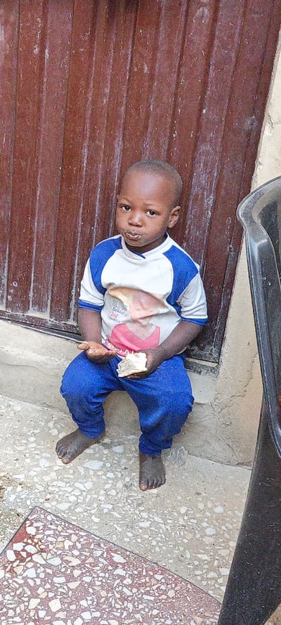The image displays a full-body shot of a young, dark-skinned child seated on a concrete surface. The child is looking directly at the viewer with a neutral, slightly pouting expression, and there is a visible white residue around their mouth, suggesting they are eating.
The child is wearing a short-sleeved t-shirt that is predominantly white with blue raglan sleeves and a blue collar. On the front of the t-shirt, there is a printed image of a person with what appears to be reddish-brown hair. Below this image, text is partially visible, which reads "HAPPY Birthday" followed by "2nd" and "2013", indicating the shirt may be a commemorative item for a second birthday from that year. The child is also wearing blue full-length pants and is barefoot, with their feet resting on the patterned ground.
In both hands, the child holds a piece of light-colored bread or pastry. The piece in their left hand appears to be smaller or crumbled, while the piece in their right hand is larger and shows signs of being torn or eaten.
The background behind the child consists of a reddish-brown vertical surface, which resembles corrugated metal or weathered wooden planks, possibly part of a door or wall. The ground beneath the child is a light-colored concrete or tiled surface mottled with various shades of gray, white, and red aggregate. To the far right of the frame, a dark, partially visible object, possibly a black plastic chair or container, is present. The overall lighting suggests the photo was taken during the day, likely outdoors.