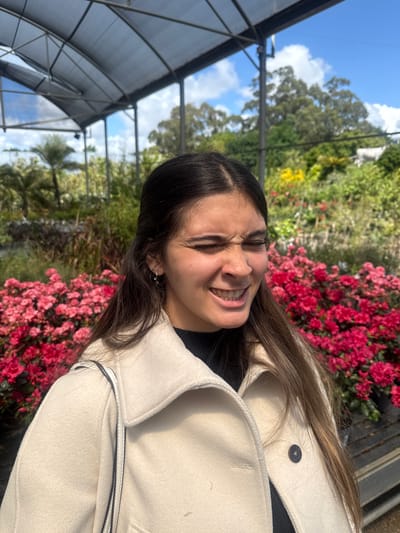 A young woman with dark hair and a light complexion is posing for a photo in what appears to be a greenhouse or nursery. Her eyes are squinted shut and she is smiling broadly, revealing her teeth. She is wearing a cream-colored coat and a black shirt underneath. Red flowering plants are visible in the background, as well as other greenery. A metallic structure, likely part of the greenhouse framework, is visible on the left. The location may be in Vila Nova de Gaia, Portugal.