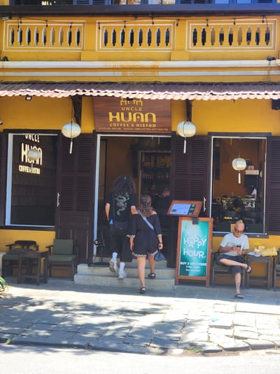 The image captures the facade of a yellow building in Hội An Ward, Vietnam, housing a coffee shop and bistro named "Uncle Huon". The building features traditional Vietnamese architectural elements with decorative yellow plasterwork along the top, dark wooden shutters on the windows and doors, and a reddish-brown tiled awning. Two white paper lanterns hang outside the establishment.

In front of the entrance, a man with long, curly dark hair and wearing a black t-shirt with a dragon graphic and dark pants is seen walking up the steps. Beside him, a woman in a dark dress with white trim is walking away from the camera. To the right, a man with a shaved head, wearing glasses, a light-colored t-shirt, and dark shorts, is seated on a chair outside the establishment, looking at his mobile phone. He is positioned next to a table with a drink and a yellow bag. A "Happy Hour" sign, advertising "BUY 2 GET 1 FREE 6 PM - 8 PM For Cocktail", stands prominently near the seated man. The ground in front of the building is paved with stone tiles, with shadows from trees dappled across it. The overall scene depicts a street-level view of a commercial establishment in a Vietnamese city.