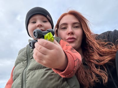 The image is a low-angle selfie featuring an adult female and a young child, positioned outdoors under a uniformly overcast sky. The adult, located on the right, has long, wavy reddish-brown hair and light eyes, with her lips slightly pursed. She is wearing a dark outer layer and an orange/rust-colored long-sleeved garment visible on her left side. The child, situated on the left and slightly forward, appears to be a toddler or preschooler, wearing a black knitted beanie with a visible tag, an olive green puffer vest with a silver zipper, and an orange/rust-colored long-sleeved top beneath. The child holds a small, green and black toy vehicle, resembling a monster truck, in their right hand, presenting it towards the camera. Both subjects are looking directly into the camera. The specific geographic location, including any city or country such as Elovo, Russia, is not discernible from the image content.