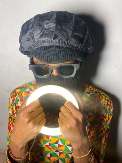An individual is depicted standing against a plain, light-colored wall in Ngleshi Amanfro, Ghana. The subject wears a black balaclava covering the entire face except for the eyes, which are obscured by black, rectangular-framed sunglasses. A crinkled black fabric cap is worn atop the balaclava. The individual is dressed in a vibrant, multi-colored shirt featuring a geometric pattern of yellow, red, green, black, and white squares and diamonds. The left wrist displays an orange bracelet, and a thin golden string bracelet is on the right wrist. Both hands hold an illuminated circular LED ring light directly in front of the subject's upper torso, emitting a bright white light.