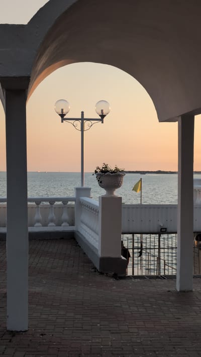 The image shows an outdoor scene in Gelendzhik, Russia, at sunset. The foreground features a walkway with a decorative white railing and a paved brick surface. A large, arched structure frames the left side of the image, casting a shadow. A lamppost with two globe-shaped lights stands prominently on the walkway.

Beyond the walkway is a body of water, likely the sea, reflecting the soft hues of the sunset. A yellow flag is visible in the distance, fluttering on a pole. A white balustrade separates the walkway from the water's edge. A decorative vase filled with flowers sits on a pedestal near the balustrade. The sky is a gradient of warm orange and pink, typical of a sunset.

In the background, a metal fence overlooks the water. The overall atmosphere is serene and peaceful, suggesting a quiet evening by the water. There is no visible text in the image. The scene evokes a sense of calm and beauty associated with sunset vistas.