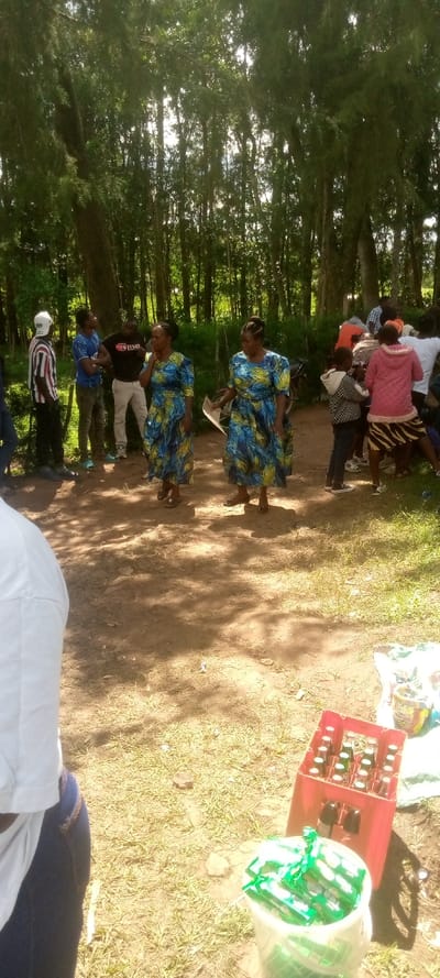 The scene takes place in a grassy clearing in a forested area in Kivaywa, Kenya. There are several people in the clearing, some are standing, others are sitting on the ground. There are two women wearing blue and yellow patterned dresses. There are several men wearing shirts, and some children in the background. A red crate of glass bottles sits on the ground next to a bucket of green, rectangular items. There are several large trees in the background. The ground is dirt and grass. The sun is shining brightly.