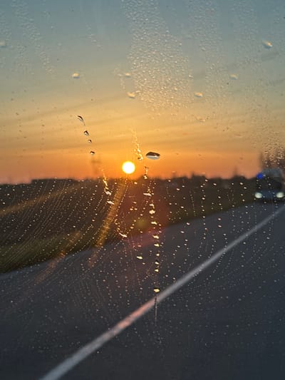 The image captures an outdoor scene at either sunrise or sunset, viewed through a transparent surface, likely a vehicle windshield, densely covered in water droplets. A bright, circular orange sun is positioned low on the horizon, slightly left of center, illuminating a sky that transitions from light blue overhead to warm orange and yellow near the horizon. A faint, dark silhouette, possibly a power pylon, is visible directly behind the sun. Below the horizon, a dark asphalt road extends into the distance, with a solid white line traversing it diagonally from the lower left to the mid-right. A narrow strip of green vegetation runs along the left side of the road. In the far distance on the right side of the road, blurred vehicle lights are visible, including two white headlights and a distinct blue flashing light. The water droplets on the viewing surface refract and reflect the sun's light, creating numerous bright points and streaks. This scene is located in the city of Marcoussis, France.