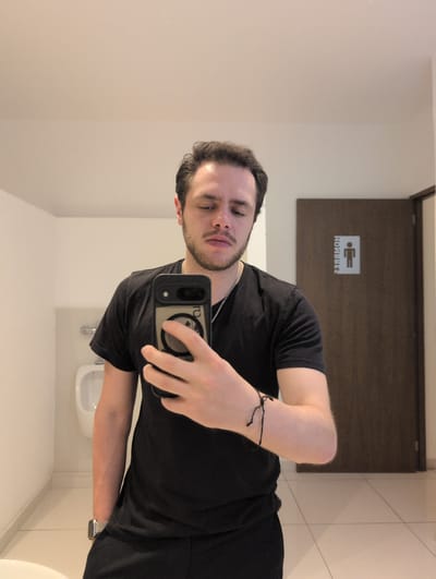 A fair-skinned man is taking a selfie in a restroom with a sign for “Hombres” above his head. He is wearing a black t-shirt, a silver necklace and a thin black bracelet on his left wrist. He is holding a smartphone in his right hand. The restroom has a white sink and white tile on the walls. The photograph was taken at an unknown location in Jesús del Monte, Mexico.