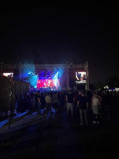 The image depicts an outdoor music concert at night. A large stage with bright blue and red lights is visible in the background, featuring large screens displaying the performers. A crowd of people is gathered in front of the stage, illuminated by the stage lights. In the foreground, a dark tent-like structure is present on the left, with people milling around. The ground appears to be asphalt, wet and reflective due to the lighting. The overall atmosphere suggests a lively event. The location is Palilula Urban Municipality, Serbia.