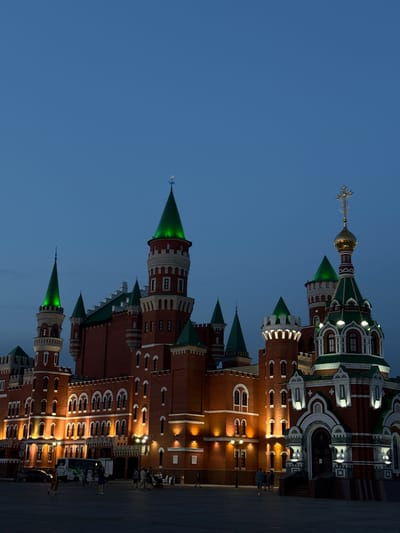 This is a vertical, medium-shot, low-angle, outdoor photograph of the National Art Gallery in Yoshkar-Ola, Russia, at dusk. The building, which resembles a medieval castle, is made of red brick and features numerous towers with green, conical roofs. Several of these towers are illuminated from within, casting a green glow. A golden-domed Orthodox church with a green roof and a cross on top is visible to the right of the castle structure. The sky is a deep twilight blue. In the foreground, a paved square is visible, with several people walking and standing. A bus and a car are parked on the left side of the square. Streetlights illuminate the building's facade.