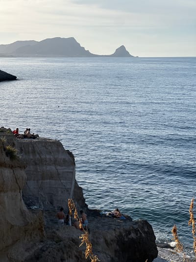 This vertical photographic image depicts a coastal scene near Sperone, Italy, under diffused sunlight, likely during late afternoon. The composition is divided by a rugged cliff face in the foreground and lower midground, with the Mediterranean Sea and distant landmasses occupying the remainder of the frame.
The foreground is dominated by a light-colored, layered rock cliff exhibiting visible strata and erosion. Sparse, dry vegetation is scattered on the cliff ledges, particularly on the upper left. In the lower right corner, out-of-focus dried plant stalks with golden hues are prominent.
On an upper, relatively flat ledge of the cliff, located on the left side of the image, four individuals are visible. They appear to be resting or sunbathing, with one person wearing a red top and others in light attire; towels or personal items are present on the rock next to them. Further down the cliff face, at the water's edge on a lower rock shelf, another group of four individuals is present. These individuals are mostly shirtless, some with towels, suggesting recent or impending swimming activity. One person is partially in the water. A white garment is draped over a rock near this group.
The sea spans the middle and upper sections of the image, displaying a deep blue color with visible ripples and glinting highlights from the sun. A single red buoy floats in the water in the mid-distance to the left of the image. Several smaller, dark specks, possibly buoys or birds, are scattered across the water's surface.
The distant horizon features two prominent landmasses silhouetted against a pale, slightly hazy sky. To the left, a wide, undulating mountain range with hints of urban development at its base appears as a dark, indistinct form. To the right, a distinctive, sharper, conical peak rises directly from the sea.
