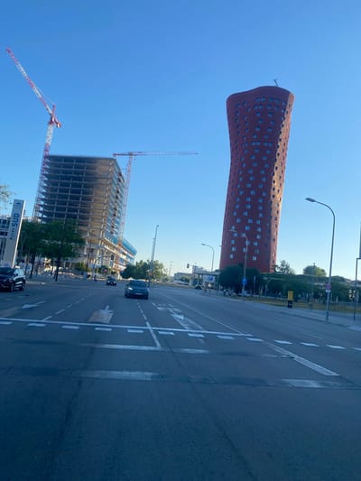 The image captures an urban scene on a bright, sunny day, likely late morning or early afternoon given the strong shadows. The perspective is from ground level, looking up at tall buildings and a wide street.

Dominating the left side of the frame is the distinctive curved, red brick facade of a skyscraper, with numerous windows. To its right and closer to the foreground, a multi-story building is under construction, its concrete framework visible, with construction cranes extending into the clear blue sky. One crane is particularly prominent in the foreground, its red and white boom angled across the frame. Another, taller crane is visible in the distance, also against the blue sky.

The street in the foreground is paved and marked with white traffic lines, including directional arrows and pedestrian crossing markings. Several cars are visible on the road, suggesting ongoing urban activity. Trees line the side of the street, interspersed with lampposts and some signage.

A sign at the bottom right of the frame is partially visible, displaying the text "ACCES SUD". The overall atmosphere is one of a modern, developing urban environment. The clear blue sky suggests fair weather. The location appears to be in a commercial or business district, possibly near a construction zone, given the presence of the building under construction and the cranes.