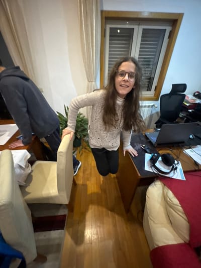 An indoor scene in Mirandela, Portugal, depicts two individuals within a furnished room. The primary subject, a young female with long brown hair and glasses, is positioned centrally, smiling directly at the camera. She wears a light-colored knitted turtleneck sweater and dark pants, and appears to be suspended mid-air with her feet off the polished wooden floor. Her left hand rests on the back of a light-colored upholstered chair, and her right hand is placed on a brown wooden desk to her right. To the left, a male figure is partially visible from the back, leaning over a separate wooden table covered with white papers. He wears a dark blue hooded sweatshirt and dark pants.
The room features a large window with a wooden frame and light-colored curtains in the background. Below the window, a white radiator is visible. To the right of the window is a black office chair. The wooden desk, where the female subject's right hand rests, holds an open black laptop, a black computer mouse, a black headset, and several white papers. In the foreground to the right, a section of a light-colored sofa or armchair draped with a red blanket is visible. A green potted plant is situated between the two tables. The floor is composed of polished wooden planks, and a striped rug is partially visible under the chair to the left. Overhead lighting casts shadows onto the floor.