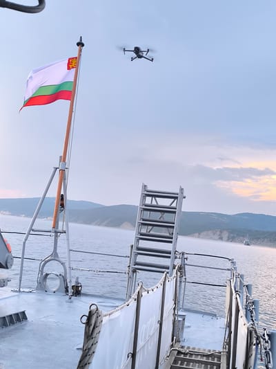 The image captures a view from the deck of a vessel at sea, likely near the coast of Emona, Bulgaria, given the presence of the Bulgarian flag. A drone is visible flying in the clear sky. In the foreground, part of the ship's deck is visible, including a ladder, railings, and a white banner. The Bulgarian flag, featuring white, green, and red stripes with a coat of arms on the white stripe, is prominently displayed on a flagpole. The background shows a coastline with hills and a distant ship on the water. The lighting suggests it is either dawn or dusk, with soft, diffused light illuminating the scene and a hint of color in the sky.