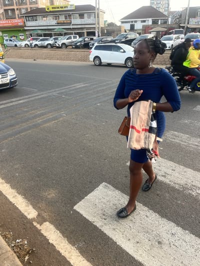 A woman in a blue dress crosses a street on a pedestrian crossing. She carries a beige bag with a red stripe and a brown purse. Cars are parked along the street, and traffic passes by. A building labeled "Overstone Park Hotel" is visible. The woman is wearing black flats and a gold bracelet. Tire tracks are present on the road. The lighting suggests it's daytime.