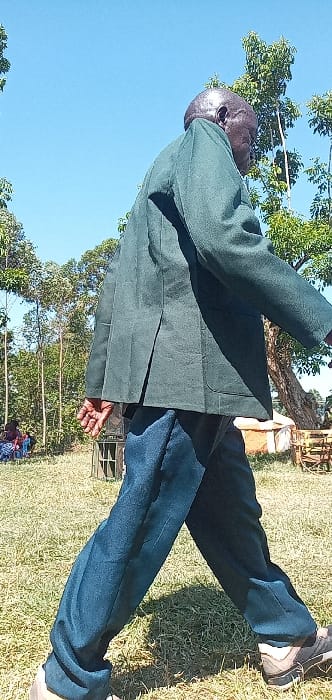 An outdoor, brightly lit, full-body shot captures an older man, likely of African descent, standing in Burundu, Kenya. He is positioned towards the left of the frame, with his body slightly angled and his bald head turned to face the right. The man is wearing a dark green jacket or coat over a dark blue shirt and dark blue trousers, paired with dark sneakers with light soles. His right hand is visible, open and relaxed.
The setting is a grassy field under a clear, bright blue sky, indicating sunny weather. Several trees, featuring green foliage, line the background, with some taller, slender trees extending upwards. The ground cover consists of green grass with some drier, brown patches. Strong shadows are cast, suggesting it is either mid-morning or early afternoon. In the distant background, faint shapes hint at other people or structures within the natural landscape. There is no visible text in the image.