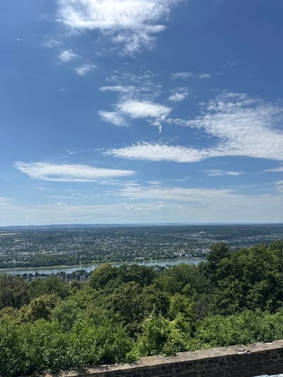 The image captures a panoramic view of Königswinter, Germany, from an elevated vantage point. In the foreground, a stone wall runs horizontally, with lush green foliage and trees obscuring much of the lower part of the scene. Beyond this, a wide river, likely the Rhine, snakes through the landscape, bordered by trees on its banks. Across the river, the city of Königswinter sprawls, a dense collection of buildings interspersed with greenery. The cityscape extends towards a distant horizon marked by rolling hills and more vegetation. The sky above is a vibrant blue, dotted with scattered white clouds, some wispy and others more defined. A small airplane is visible in the upper portion of the sky. The overall impression is one of a sunny day with a clear view of the city and its natural surroundings.