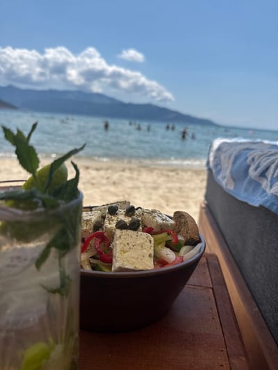 A close-up, shallow depth of field shot shows a Greek salad and a refreshing drink on a wooden table, with the beach and sea in the background. The salad, in a dark bowl, features large cubes of feta cheese, capers, sliced red peppers, and a piece of bread. The drink, in a clear glass to the left, contains ice, mint leaves, and a lime wedge. The blurred background reveals a sandy beach, the turquoise sea with several people swimming, and distant hills under a bright blue sky with scattered clouds. The scene suggests a relaxed, seaside dining experience in Keramoti, Greece.