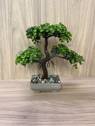 A single, artificial bonsai tree is centrally placed on a light brown wooden surface, against a backdrop of vertical wooden planks. The tree has a dark brown trunk and branches, adorned with numerous small, bright green leaves. The base of the tree is a light gray rectangular pot filled with small, mixed-color pebbles. The scene is static, with no people or animals present. The lighting is even, casting soft shadows. The location is not specified in the image.