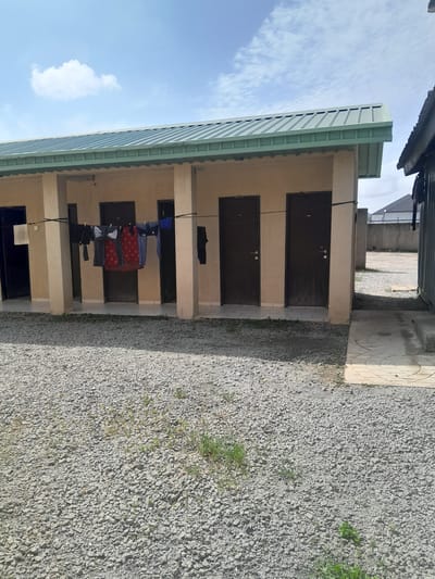 The image shows a row of rooms or units with light beige walls and dark brown doors. A clothesline is strung across the front of some of the units, with clothes hanging on it to dry. The ground in front of the units is covered in small gray gravel, with some patches of green grass. The building has a green, corrugated metal roof. In the background, there is a concrete wall with a fence on top, and a cloudy blue sky. The location is identified as Dam Camp, Nigeria.