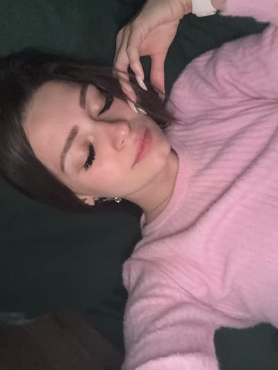 A woman with long dark hair, wearing a pink sweater and a white wristband, is lying down with her eyes closed. She has long eyelashes and manicured nails. Her face is slightly tilted. She is wearing an earring on one ear. The background appears to be a dark green, possibly a couch or cushion. The location is not discernible from this image.